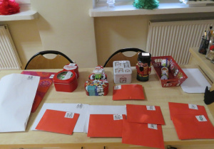 Na ławce znajdują się czerwone koperty i puszki świąteczne z zadaniami do wykonania.
