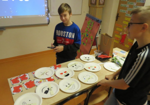 Dwóch uczniów dopasowuje przymiotniki z nazwami emocji do białych talerzy, na których narysowane są emotikony.